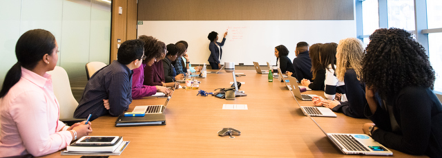 A group of people sitting at a large conference room table, looking at a person at a whiteboard